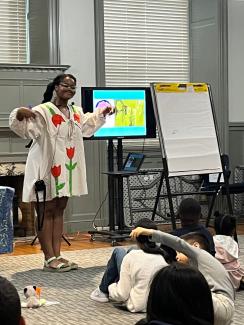 Author Oge Mora standing next to a screen speaking to a room of children