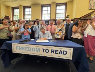Governor Dan McKee seated at a table while signing the Freedom to Read bill. Excited onlookers and the bill's supporters are clapping as they stand behind him.